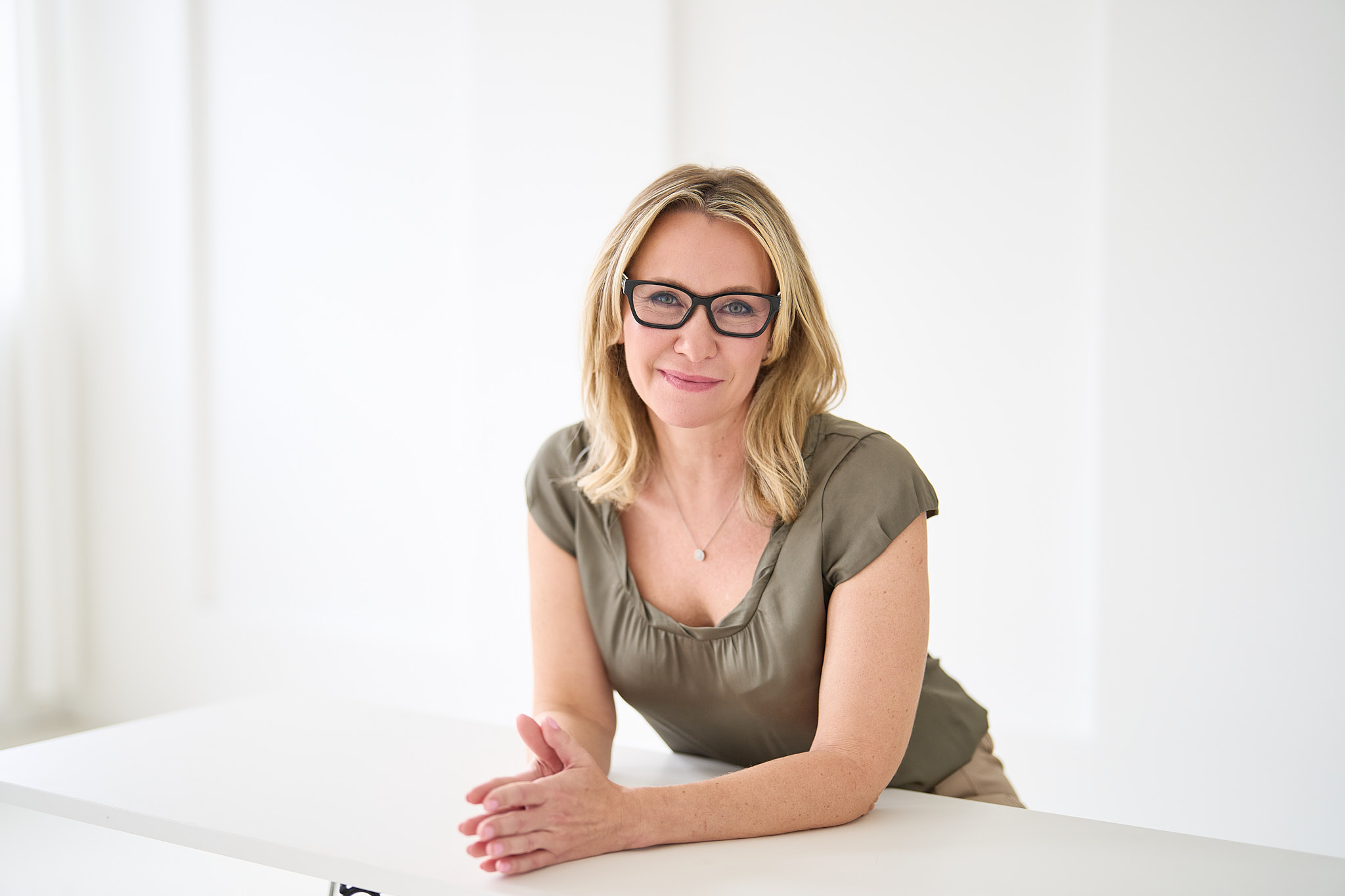 Blonde Frau mit Brille und olivgruener Bluse laechelt freundlich am weissen Tisch
